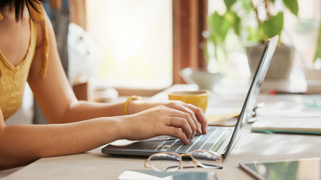 A woman in a yellow top types on a laptop in a sunlit room. A potted plant, glasses, and a smartphone are on the desk.